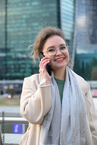 A young woman of European appearance speaks on the phone outdoors in the afternoon against the backdrop of the skyscrapers of Moscow City