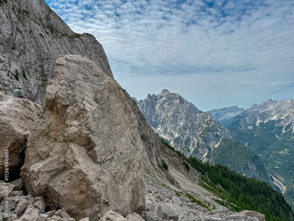 Hiker man sitting on massive rock formation with panoramic view of rugged mountain ridge Prisank in majestic Julian Alps. Adventure and freedom in Triglav National Park, Slovenia, Slovenian Alps Hiker man sitting on massive rock formation with panoramic view of rugged mountain ridge Prisank in majestic Julian Alps. Adventure and freedom in Triglav National Park, Slovenia, Slovenian Alps