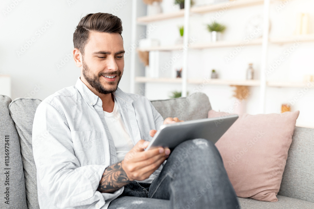 Relaxed millennial man resting on sofa with digital tablet, free space. Positive guy browsing social networks or shopping online while sitting in living room interior Relaxed millennial man resting on sofa with digital tablet, free space. Positive guy browsing social networks or shopping online while sitting in living room interior