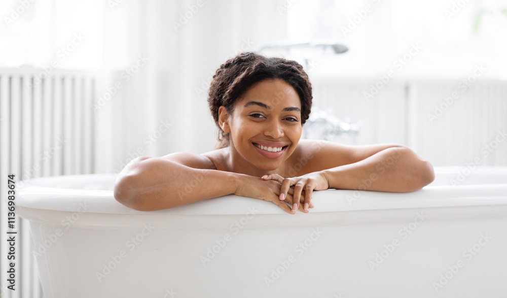 A pretty African American woman smiles while relaxing in a bright bathroom. She enjoys a moment of self-care at home, embracing tranquility and pampering with skincare. A pretty African American woman smiles while relaxing in a bright bathroom. She enjoys a moment of self-care at home, embracing tranquility and pampering with skincare.