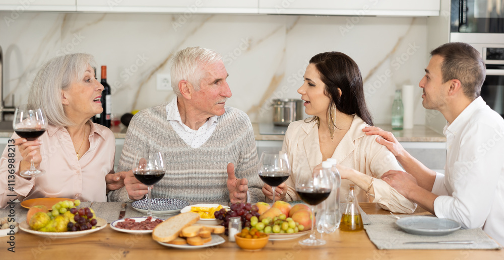 Cozy dinner scene while elderly parents enjoying wine and meal with adult daughter and son-in-law in well-lit home kitchen.. Cozy dinner scene while elderly parents enjoying wine and meal with adult daughter and son-in-law in well-lit home kitchen..