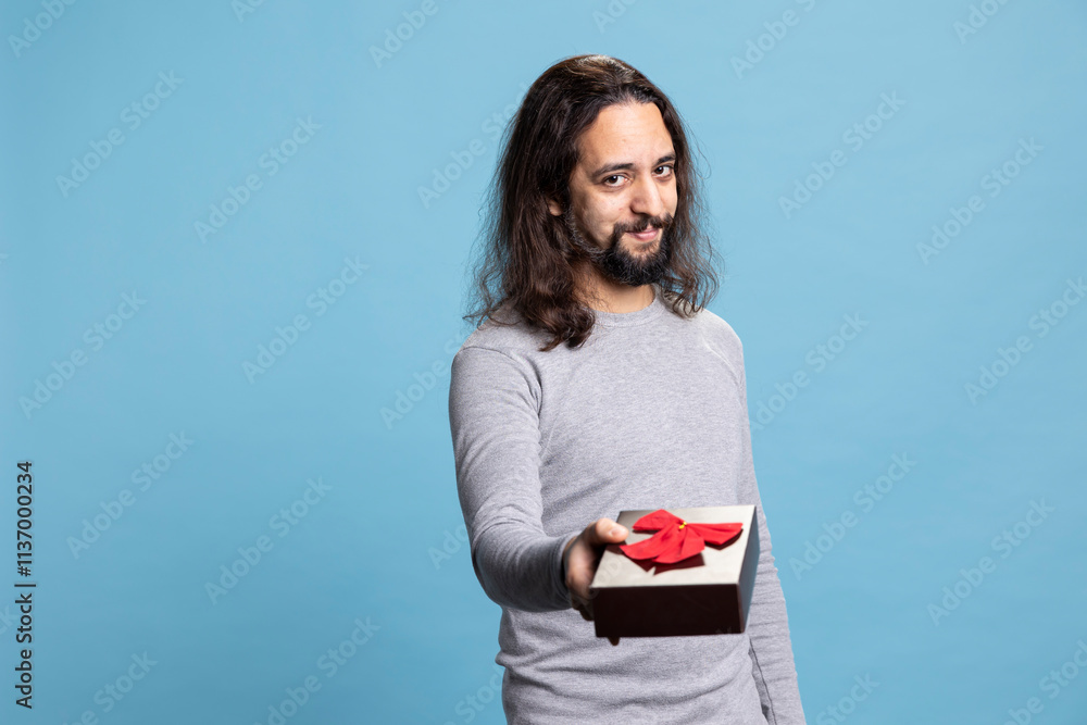 Sweet affectionate guy offering a gift in a cute box with ribbon on camera, giving presents for Christmas or anniversary against blue background. Person in casual wear showing gratitude and love.