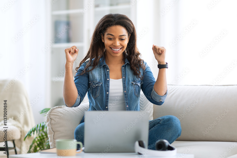 A young black woman is sitting on a couch at home, smiling and celebrating. She raises her arms in excitement, clearly enjoying a moment of success as she works on her laptop in a bright room. A young black woman is sitting on a couch at home, smiling and celebrating. She raises her arms in excitement, clearly enjoying a moment of success as she works on her laptop in a bright room.