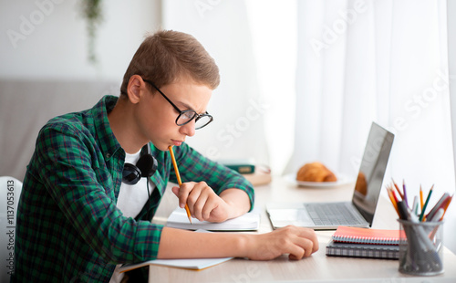 Intelligent schooler working hard on homework, sitting at desk at home and writing essay, online education concept