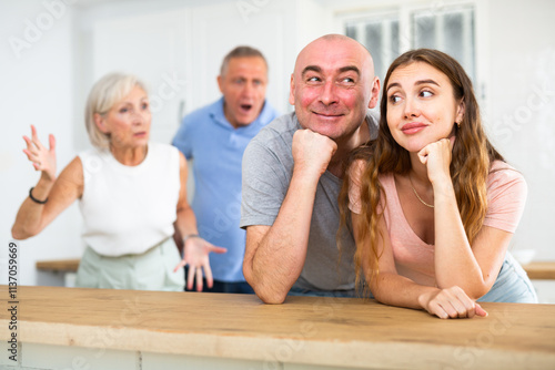 Portrait of a happy married couple in a home kitchen, not paying attention to adult family members who reprimand them photo