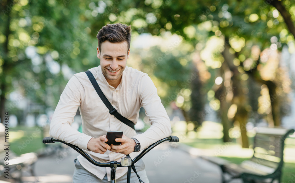 Social networks outdoors. Smiling attractive guy in shirt and trousers sits on bicycle in park and looks at smartphone, free space Social networks outdoors. Smiling attractive guy in shirt and trousers sits on bicycle in park and looks at smartphone, free space