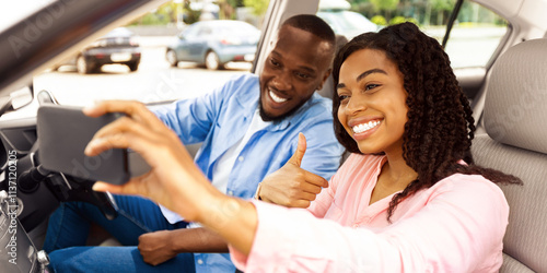 Beautiful young African American couple taking selfie inside car, using mobile phone, showing thumbs up sign gesture. Happy black man and woman going on vacation, taking self photo, buying new auto