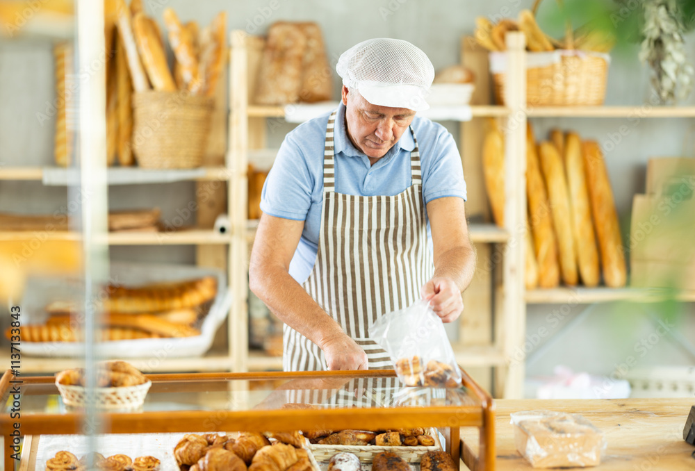 Elderly man seller in apron putting pastries in plastic bag in bakery Elderly man seller in apron putting pastries in plastic bag in bakery