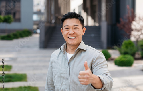 Portrait of positive asian man standing outdoors and gesturing thumb up, walking in urban city area, looking and smiling at camera, free copy space photo