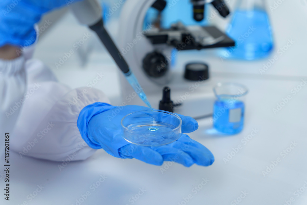 Close up of scientist's hand in blue gloves dropping blue liquid on petri dish using pipette, with microscope and beakers in background Close up of scientist's hand in blue gloves dropping blue liquid on petri dish using pipette, with microscope and beakers in background