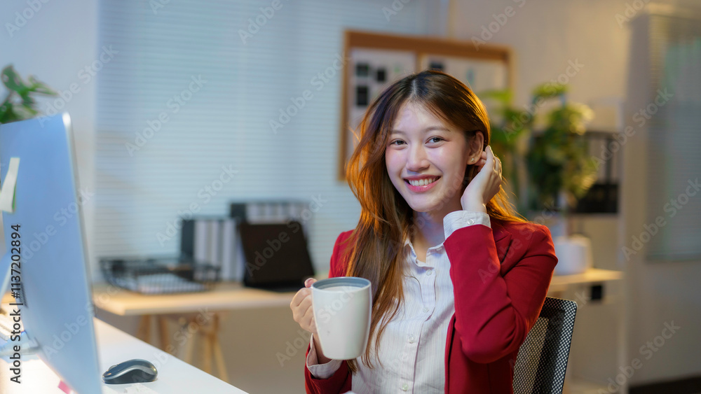 Young office worker smiling while holding a coffee mug, immersed in late night tasks at her desk, surrounded by modern technology Young office worker smiling while holding a coffee mug, immersed in late night tasks at her desk, surrounded by modern technology