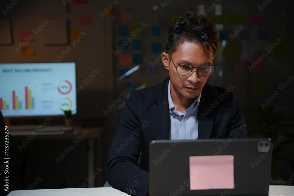 Young businessman working late at night in office using laptop, with graphs on a monitor in the background Young businessman working late at night in office using laptop, with graphs on a monitor in the background