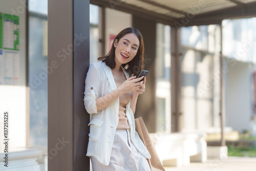Free Download Happy businesswoman waiting at the bus stop, using her smartphone while holding a shopping bag, enjoying urban life and convenience - GFXLoad
