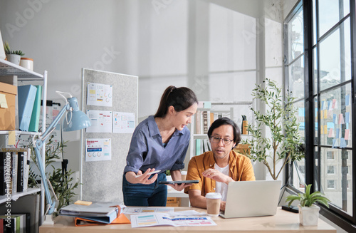 Two young Asian startup company partners discuss and brainstorm in casual small business office, marketing planning jobs on a laptop, and freelance work as E-commerce entrepreneur and coworker team.