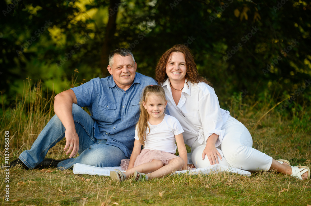 Middle aged parents with daughter sitting on grass during picnic enjoying time together smiling happily. Happy family concept, vacation, family values. Middle aged parents with daughter sitting on grass during picnic enjoying time together smiling happily. Happy family concept, vacation, family values.
