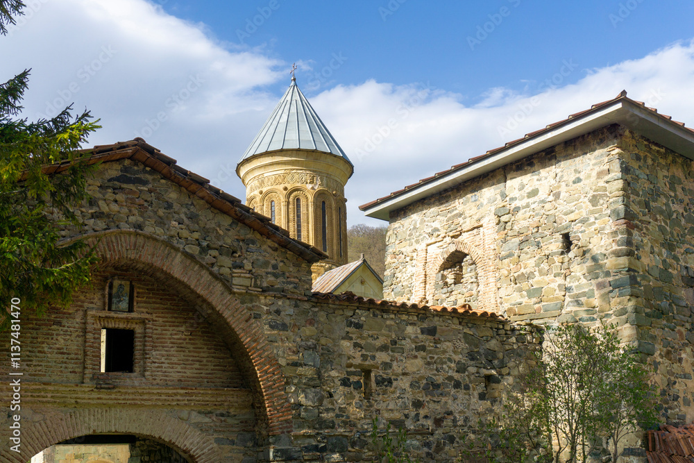 Entrance to the courtyard of the Kvatakhevi monastery. Stone walls, metal gates, arch, tower, dome of the church. Bright blue sky with clouds. Entrance to the courtyard of the Kvatakhevi monastery. Stone walls, metal gates, arch, tower, dome of the church. Bright blue sky with clouds.