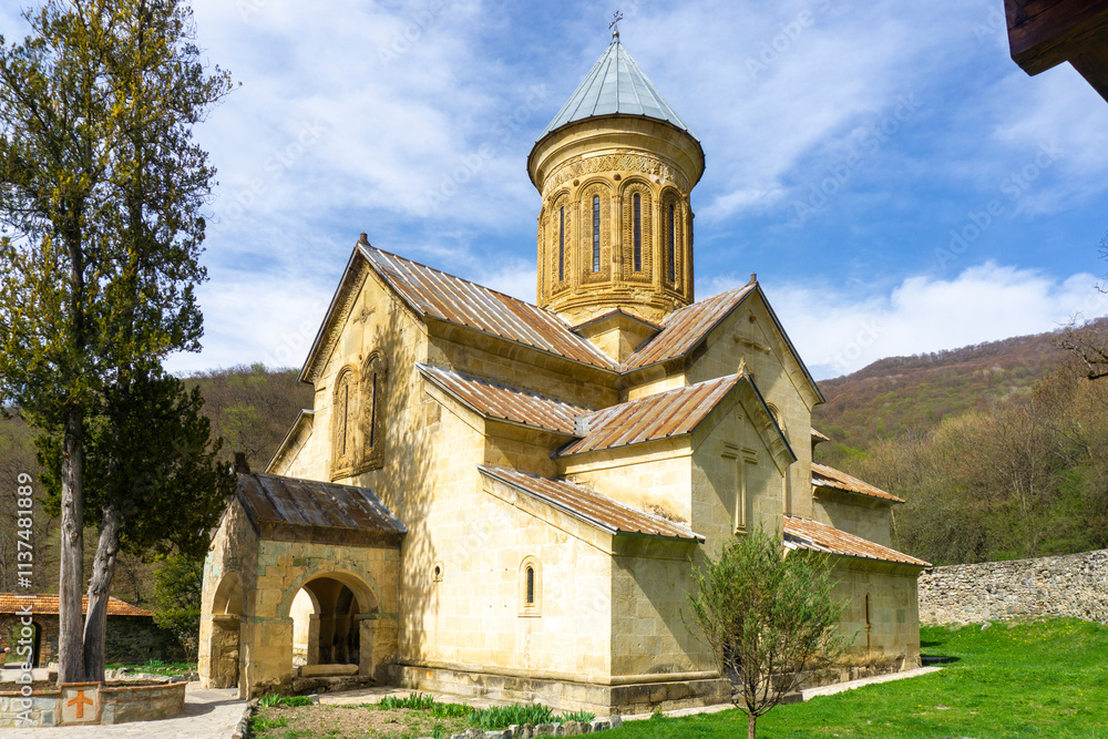 Church in the courtyard of the Kvatakhevi monastery. Stone walls, tiled roof. Stone paths, green grass, bright blue sky. Church in the courtyard of the Kvatakhevi monastery. Stone walls, tiled roof. Stone paths, green grass, bright blue sky.
