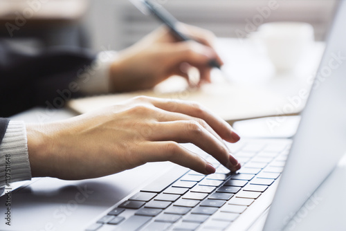 Female hands in sharp focus typing on a modern laptop keyboard, office environment in soft blur behind