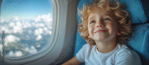 A 10-year-old Caucasian boy is sitting on a plane, wearing a white T-shirt, smiling from the side, with blue seats, blue sky and white clouds outside the window.