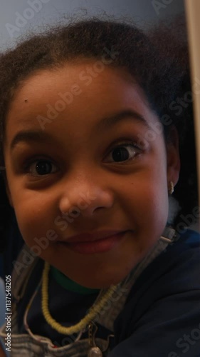Vertical of little biracial girl opening door while playing hide-and-seek game at cozy home in evening and finding player behind camera gazing with contentment