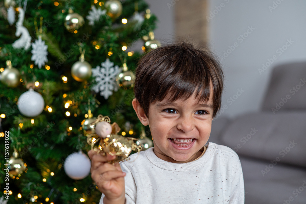 A young boy is holding a gold ornament in his hand, smiling and looking at the camera. The scene takes place in a living room, with a Christmas tree in the background A young boy is holding a gold ornament in his hand, smiling and looking at the camera. The scene takes place in a living room, with a Christmas tree in the background