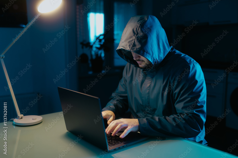 Man in Hoodie Working on Laptop in Dimly Lit Kitchen at Home Man in Hoodie Working on Laptop in Dimly Lit Kitchen at Home