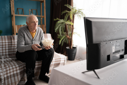 Elderly man watching TV and eating popcorn on sofa. Retired grandfather with remote control choosing interesting movie photo