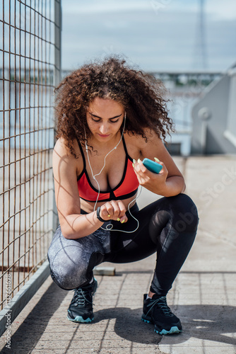 Young athletic woman with smartphone and earbuds crouching outdoors looking at wristwatch photo