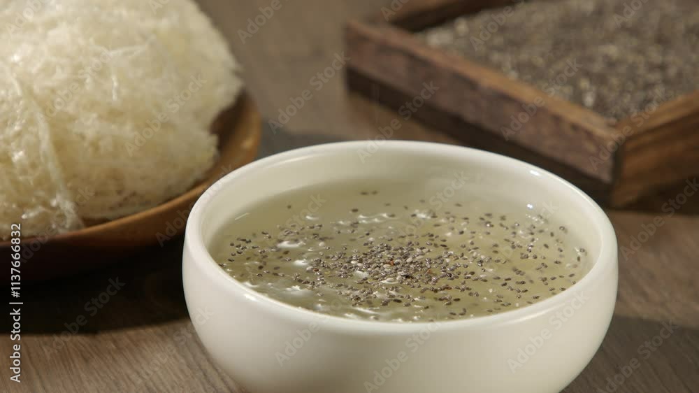 A frontal view of a model hand is sprinkling chia seeds on bird's nest soup, placed on a wooden surface next to raw swallow nests and a chia seeds tray; typical food enhances the body's resistance.