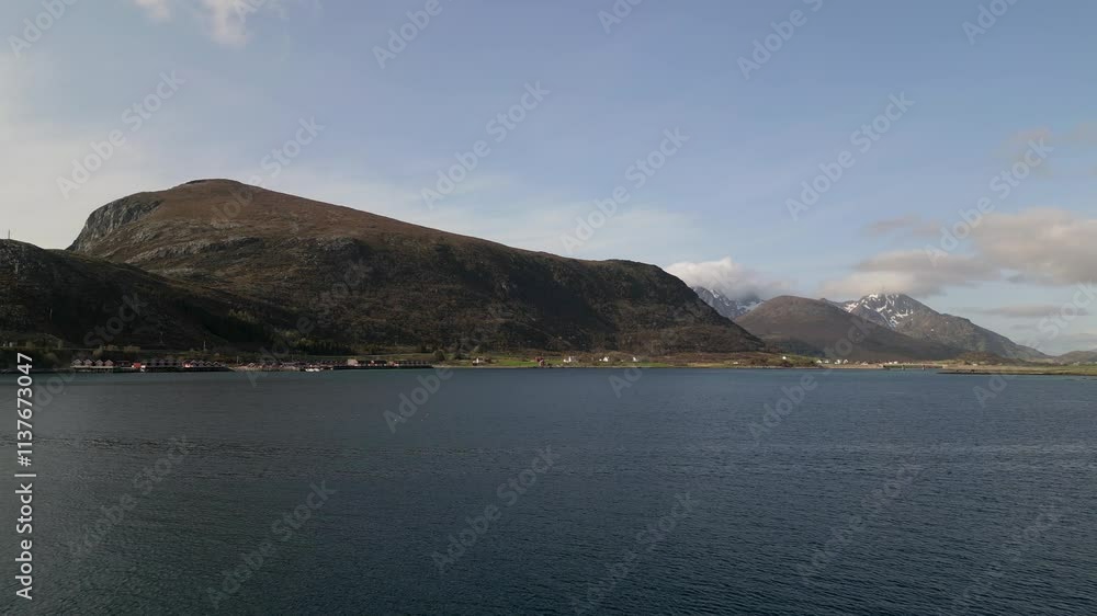 From the shores of offersøy island, Offersøykammen mountain features slopes descending to a tranquil bay, framed by distant snow-capped peaks in the Lofoten archipelago, drone flying forward From the shores of offersøy island, Offersøykammen mountain features slopes descending to a tranquil bay, framed by distant snow-capped peaks in the Lofoten archipelago, drone flying forward