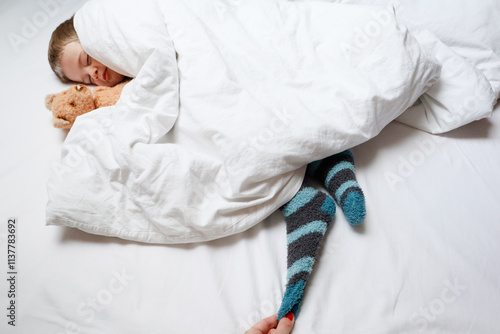 A child with a teddy bear is sleeping under a white blanket photo