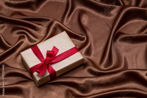 Gift box wrapped in kraft paper with red bow on silky wavy brown fabric background photo