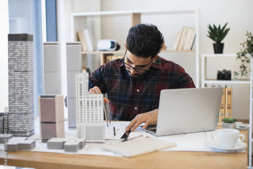 Indian male architect drawing modern city plan with pencil and paper. Young professional working on architectural project with scale models and laptop in studio. Indian male architect drawing modern city plan with pencil and paper. Young professional working on architectural project with scale models and laptop in studio.