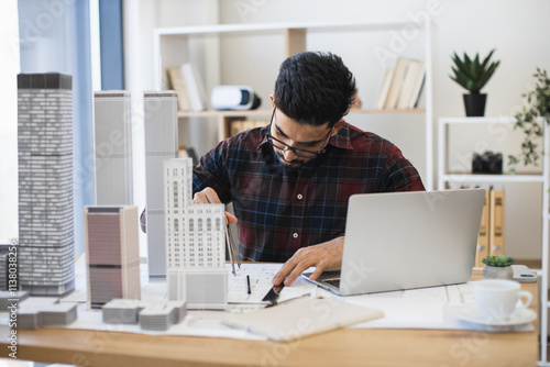 Indian male architect drawing modern city plan with pencil and paper. Young professional working on architectural project with scale models and laptop in studio.