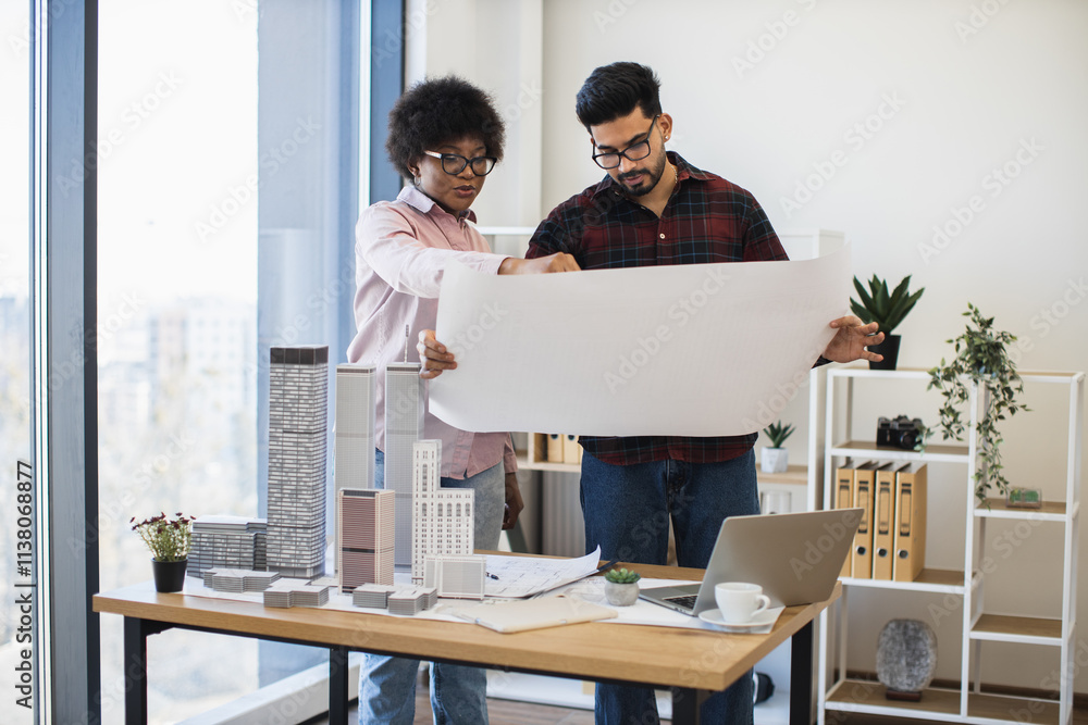 Indian man and African woman architects reviewing blueprint for modern city development. Creative office setting with desk models and laptop. Indian man and African woman architects reviewing blueprint for modern city development. Creative office setting with desk models and laptop.
