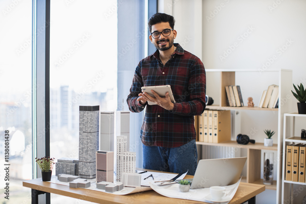 Fototapeta premium Indian male architect develops modern city project using digital tablet in creative workspace. Tall building models on desk symbolize innovative urban design. Age group young adult to middle-aged.