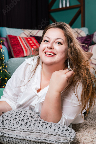 A portrait of a beautiful,chubby  woman with a sweet smile against a New Year's backdrop. A buxom woman in a white knitted sweater with an open neckline poses against a backdrop of Christmas lights.