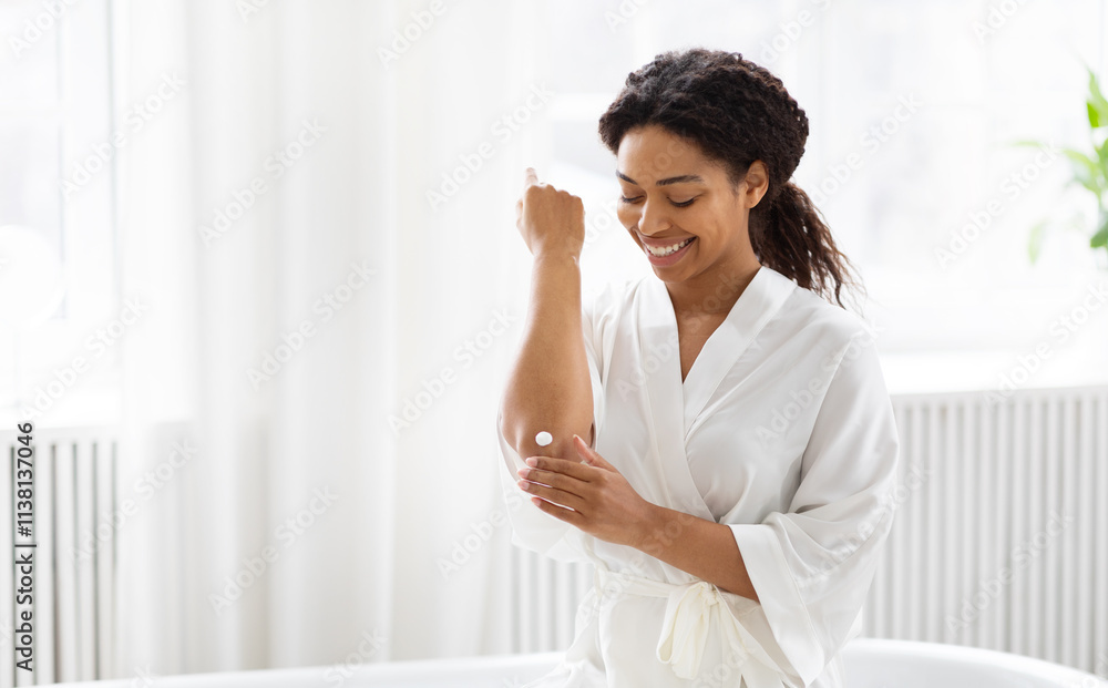 A millennial African American woman applies skin care cream to her arm, smiling softly in a bright bathroom. She enjoys a moment of pampering and self-care in her cozy space. A millennial African American woman applies skin care cream to her arm, smiling softly in a bright bathroom. She enjoys a moment of pampering and self-care in her cozy space.