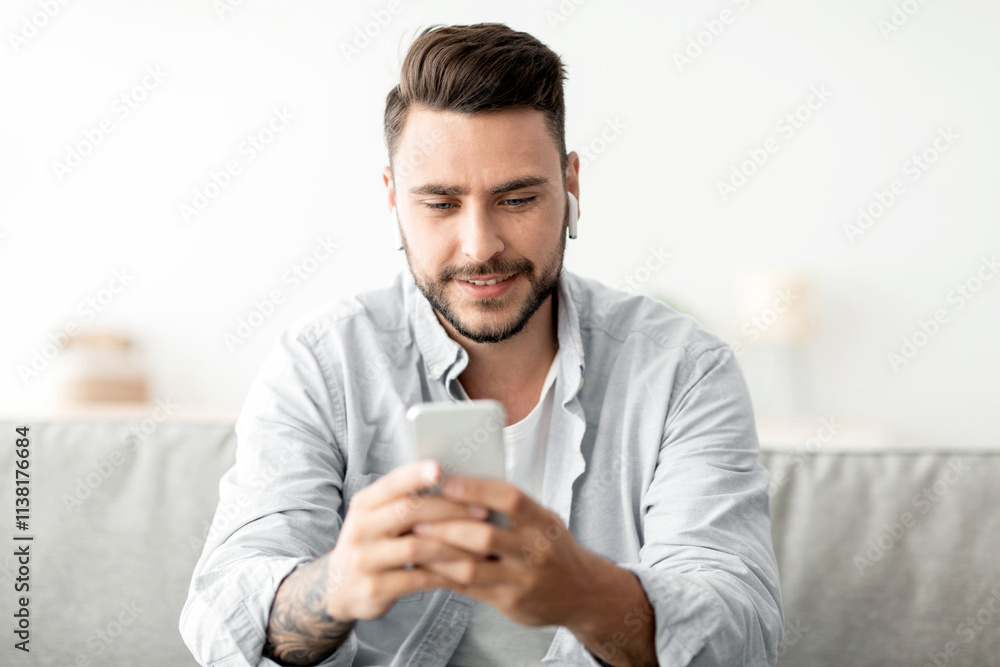 Young man with earbuds using smartphone, browsing internet and listening to music online, having video call while sitting on couch at home. Cellphone application concept Young man with earbuds using smartphone, browsing internet and listening to music online, having video call while sitting on couch at home. Cellphone application concept