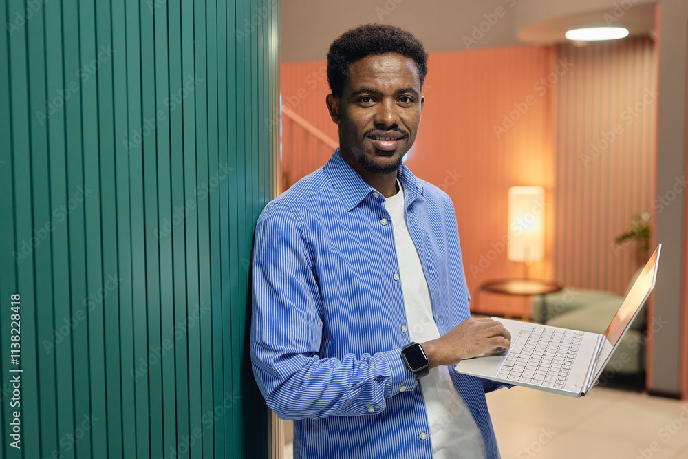 Fototapeta premium Medium portrait shot of positive African American male employee in blue shirt outfit for office holding laptop smiling at camera while standing against wall in workplace, copy space