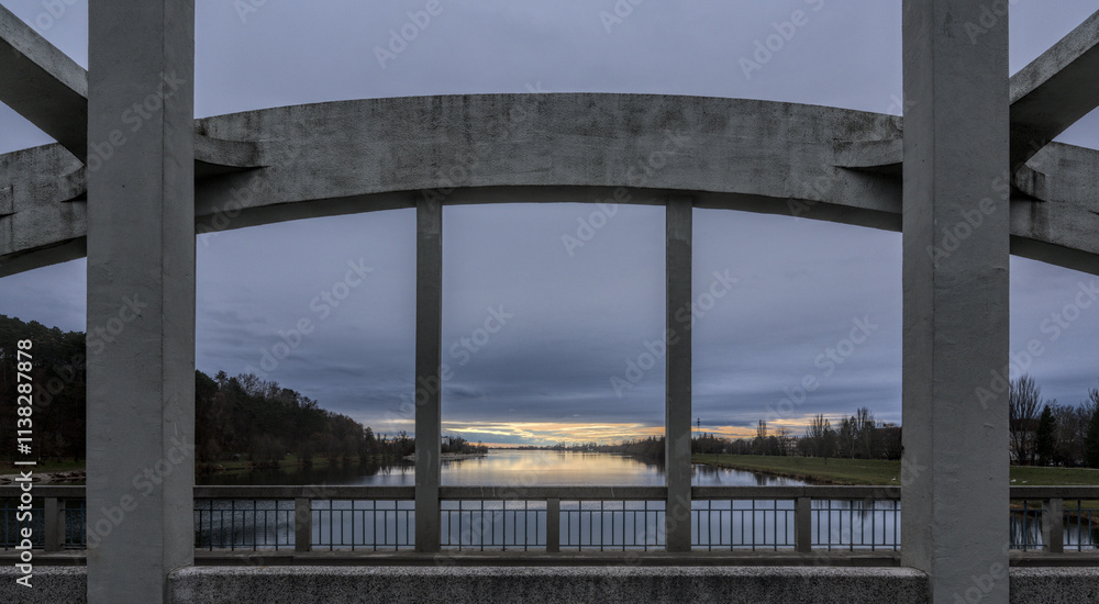 A concrete bridge arch framing a calm river, with a view of the sunset reflecting on the water under a cloudy sky A concrete bridge arch framing a calm river, with a view of the sunset reflecting on the water under a cloudy sky