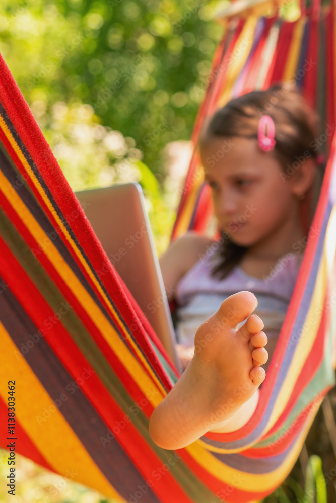 Beautiful young and happy girl lying in a hammock with laptop in a garden against green background. Freelace, remote work concept.Selective focus on bare feet. Vertical image. Beautiful young and happy girl lying in a hammock with laptop in a garden against green background. Freelace, remote work concept.Selective focus on bare feet. Vertical image.