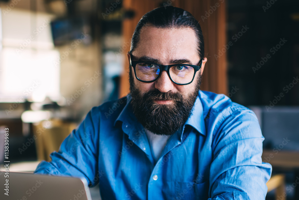 Half length portrait of serious bearded male in eyewear concentrated on remote job looking at camera, concentrated mature man 20s freelancer using laptop computer for online research in cafe Half length portrait of serious bearded male in eyewear concentrated on remote job looking at camera, concentrated mature man 20s freelancer using laptop computer for online research in cafe