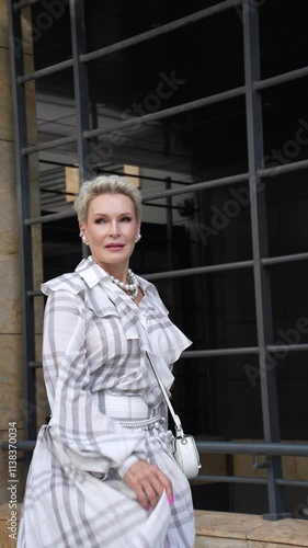 Fashionable Mature Woman with Short Hair Wearing Long Checkered White Dress, Accessories And Trendy Outfit, Walks City Streets with Confidence and Elegance. Stylish Middle Aged Fashion Model Outdoors