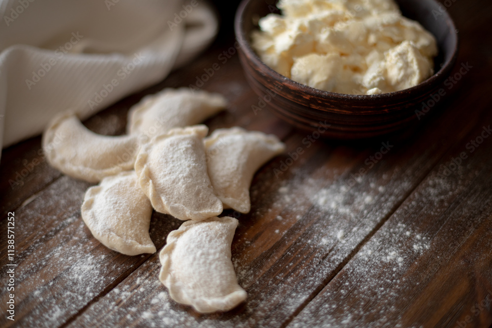 Fototapeta premium Fresh dumplings (varenyky) filled with cottage cheese, arranged on parchment paper alongside a bowl of cottage cheese and a white kitchen towel. The wooden background and flour-dusted details 