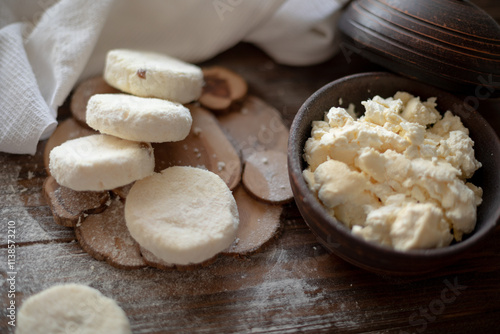 Raw cottage cheese pancakes (syrniki) with raisins, dusted with flour, arranged on a rustic wooden board alongside a dark wooden bowl filled with fresh cottage cheese. A cozy kitchen atmosphere 