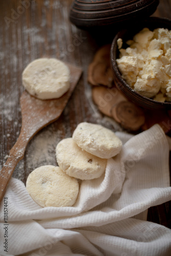 Round cottage cheese pancakes (syrniki) dusted with flour, placed on a wooden spoon and arranged next to a rustic linen kitchen towel. The warm, vintage-style kitchen background highlights a cozy atmo