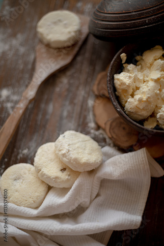 Round cottage cheese pancakes (syrniki) dusted with flour, placed on a wooden spoon and arranged next to a rustic linen kitchen towel. The warm, vintage-style kitchen background highlights a cozy atmo