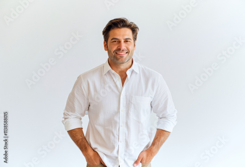 A commercial portrait photo of a model on a white studio background:  a man in a white shirt and tan pants standing with his hands in his pockets, smiling against a white background.