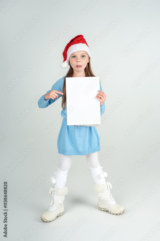Cute surprised girl in Santa Claus hat with a white blank paper sheet, copy space. Christmas and New Year sale concept. Mockup for advertising. Cute surprised girl in Santa Claus hat with a white blank paper sheet, copy space. Christmas and New Year sale concept. Mockup for advertising.